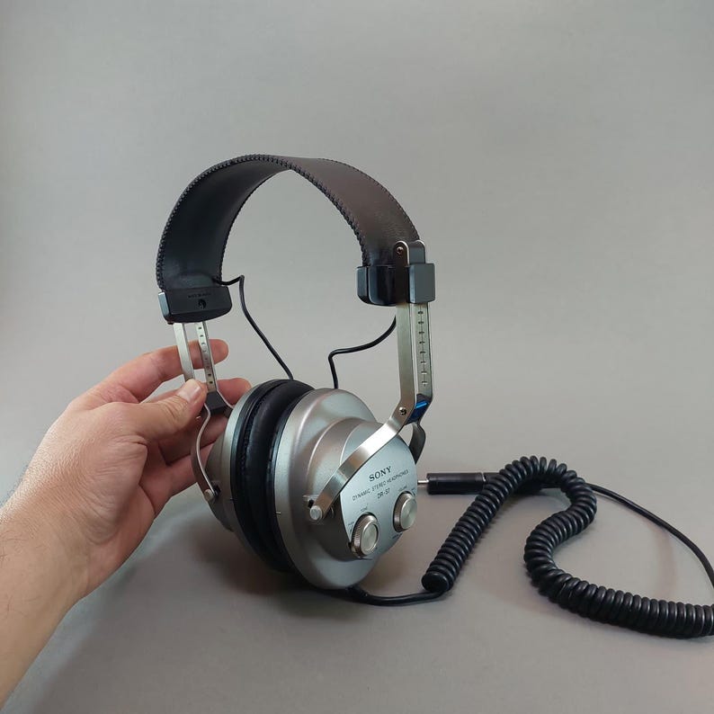 K&ouml;nnte beinhalten: Dynamischer Stereo-Kopfh&ouml;rer Sony DR-37 mit silbernen Ohrmuscheln, schwarzen gepolsterten Ohrpolstern und einem verstellbaren schwarzen Kopfb&uuml;gel. Das Ger&auml;t verf&uuml;gt &uuml;ber ein schwarzes Spiralkabel und silberne Metallb&uuml;gel mit sichtbaren Markierungen.