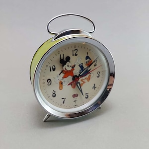 May include: A vintage alarm clock with a silver frame and a light green back. The clock face features Mickey Mouse and Donald Duck. The clock has a silver handle and legs. The hands are black and red.