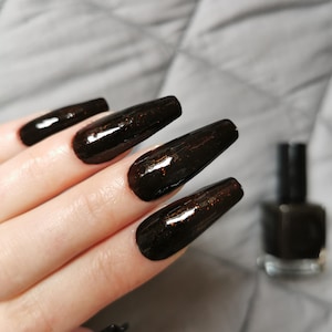 May include: Close-up of long, dark brown nails with a shimmery finish. The nails are painted with a black nail polish with a subtle copper shimmer. The bottle of nail polish is visible in the background.