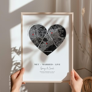 Puede incluir: Una impresión enmarcada con un diseño de mapa en forma de corazón. El mapa está dividido en tres secciones, con marcadores rojos en forma de corazón. Se muestra el texto "MET - MARRIED - LIVE", junto con nombres y fechas. El marco es de color madera clara.