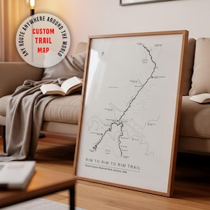 May include: A framed custom trail map print. The map is black and white, depicting the Rim to Rim Trail in Grand Canyon National Park, Arizona, USA. The frame is a light wood color. Text on the print reads "Rim to Rim to Rim Trail". A circular graphic reads "Any Route Anywhere Around The World".