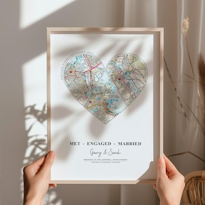 Puede incluir: Una impresión enmarcada con un diseño de mapa en forma de corazón. El mapa está dividido en tres secciones, con el texto "MET - ENGAGED - MARRIED" y los nombres "Garry & Sarah" debajo. La impresión incluye fechas y lugares.