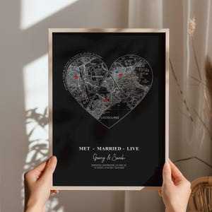 Puede incluir: Impresión enmarcada de un mapa en blanco y negro en forma de corazón, con tres ubicaciones marcadas con corazones rojos. La impresión incluye el texto "MET - MARRIED - LIVE" y nombres, con fechas y ubicaciones debajo. El marco es de color madera clara.