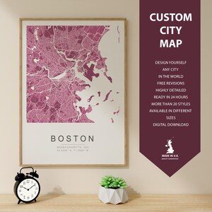 May include: Framed map print of Boston, Massachusetts, in shades of pink and white. The map is labeled "BOSTON" with coordinates. A banner on the right reads "CUSTOM CITY MAP" and lists features. A small black alarm clock and a succulent plant are in the foreground.
