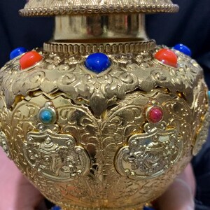 May include: Ornate gold-colored decorative vessel with intricate embossed patterns and colorful gemstone accents. The vessel features red, blue, and turquoise stones. The design includes a detailed lid and base, showcasing a blend of artistry and craftsmanship.