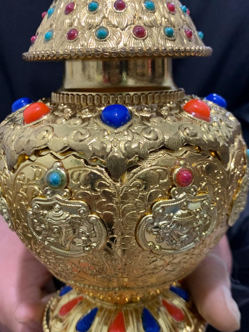 May include: Ornate gold-colored decorative vessel with intricate embossed patterns and colorful gemstone accents. The vessel features red, blue, and turquoise stones. The design includes a detailed lid and base, showcasing a blend of artistry and craftsmanship.