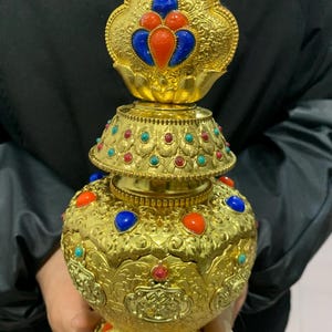 May include: Ornate gold-colored vessel with intricate patterns and colorful gemstone accents. The vessel features red, blue, and turquoise stones, and a decorative top. The design is detailed and the overall aesthetic is opulent.
