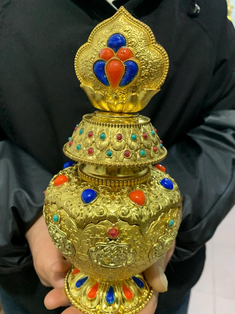 May include: Ornate gold-colored vessel with intricate patterns and colorful gemstone accents. The vessel features red, blue, and turquoise stones, and a decorative top. The design is detailed and the overall aesthetic is opulent.