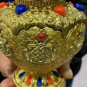 May include: Ornate gold-colored decorative vase with intricate embossed patterns. The vase is adorned with red, blue, and turquoise gemstones. The base and rim feature a repeating pattern of red and blue accents, creating a striking visual contrast.