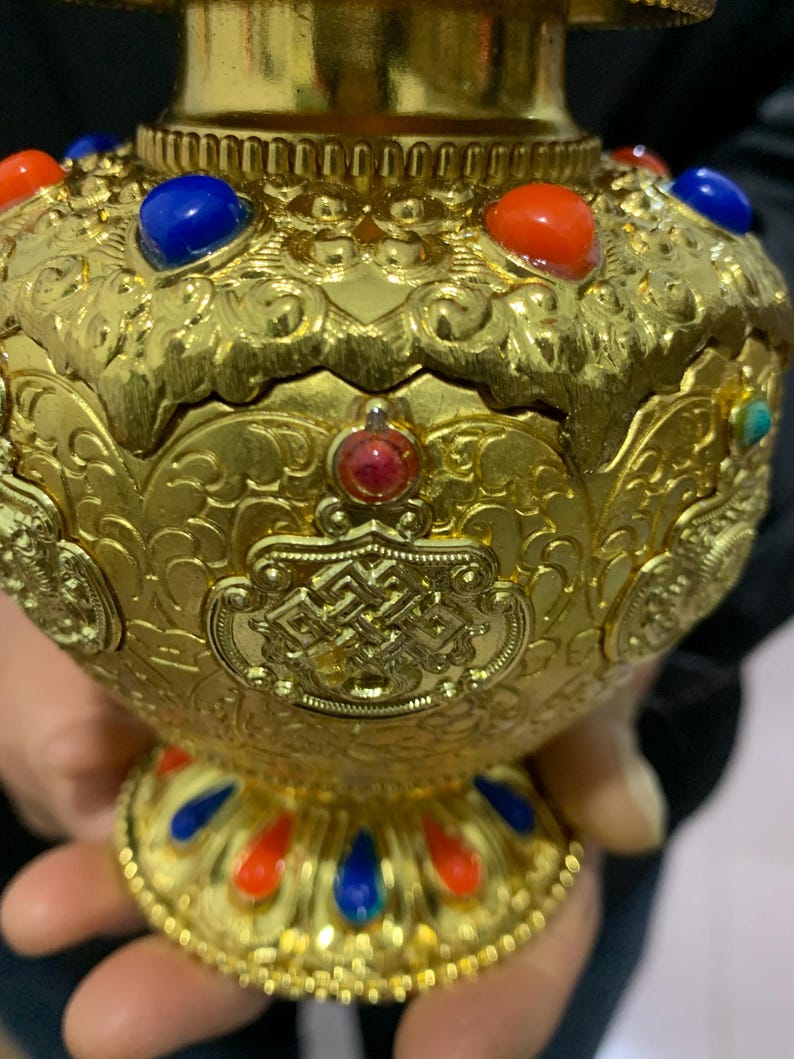 May include: Ornate gold-colored decorative vase with intricate embossed patterns. The vase is adorned with red, blue, and turquoise gemstones. The base and rim feature a repeating pattern of red and blue accents, creating a striking visual contrast.