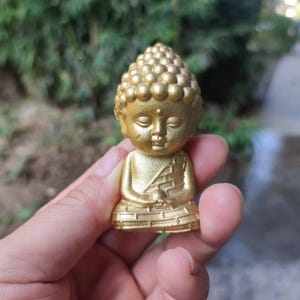 May include: A small, gold-coloured Buddha statue in a seated meditation pose. The statue has a detailed head with a textured bun and closed eyes. The figure is holding its hands in a prayer position. The statue is approximately 7.6 cm tall.