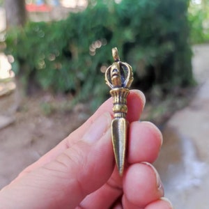May include: A bronze-colored pendant with a crown-like top and a dagger-shaped bottom. The pendant is held in a hand against a blurred green background. The pendant is likely a religious or spiritual item.