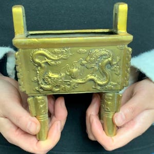 Puede incluir: Un quemador de incienso rectangular de color dorado con un diseño en relieve de dragón. El quemador tiene dos asas y tres patas decorativas. El dragón está intrincadamente detallado, y el diseño general sugiere una estética asiática tradicional.