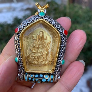May include: A pendant featuring a central gold-toned figure of a deity encased in a shield-shaped frame with intricate silver-toned metalwork. The frame is accented with small red and turquoise stones and a base cylinder with a black and blue pattern.