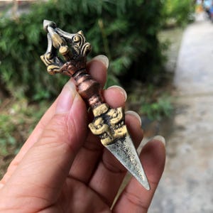 May include: A bronze-colored Vajra, a ritual object, held in a hand. The Vajra features intricate detailing with a pointed end and ornate, decorative elements. The object has a weathered appearance, suggesting age or use.