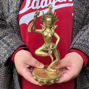 Puede incluir: Una estatua dorada de una figura danzante, probablemente una deidad, con detalles intrincados. La figura está adornada con una corona y joyas, sosteniendo una tela en una mano. Se encuentra sobre una base redonda y ornamentada. La estatua es sostenida por una persona que lleva una camisa roja y una chaqueta de tweed.