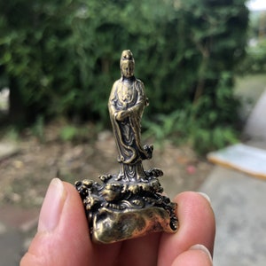 May include: A small, bronze-colored statue of a standing figure with flowing robes. The figure is on a base with detailed wave and rock formations. The statue has intricate details and a weathered appearance, suggesting an antique or vintage piece.