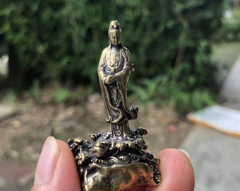 May include: A small, bronze-colored statue of a standing figure with flowing robes. The figure is on a base with detailed wave and rock formations. The statue has intricate details and a weathered appearance, suggesting an antique or vintage piece.