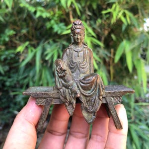 以下が含まれることがあります： 小さなベンチに座っている、おそらく神のブロンズ像。 像は流れるようなローブをまとい、装飾的な頭飾りをしています。 像は風化した外観をしており、古さやヴィンテージ感を醸し出しています。