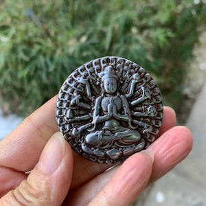 Pode incluir: Um pingente circular cinza escuro com uma representação detalhada de uma divindade sentada com vários braços. A figura está em uma pose meditativa, com as mãos juntas em oração. O pingente tem uma borda ornamentada e texturizada e um pequeno orifício na parte superior para pendurar.