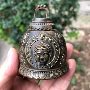 May include: Bronze-colored bell with a loop handle. The bell features an embossed image of Buddha's face within a circular frame, surrounded by floral and geometric patterns. The bell's surface has a weathered appearance.