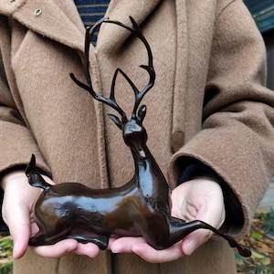 Könnte beinhalten: Eine dunkel bronzefarbene Skulptur eines Hirsches in liegender Position. Der Hirsch hat ein langes, geschwungenes Geweih, das nach oben ragt, einen schlanken Hals und eine glatte, polierte Oberfläche. Die Figur ist mit unter den Körper gezogenen Beinen dargestellt.