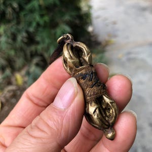May include: A small, antique brass vajra, a ritual object, is held in a hand. The vajra has a detailed design with a central grip wrapped in a dark material. The ends feature ornate, symmetrical patterns. A small loop is at the top.
