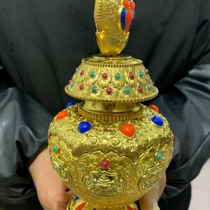 May include: Ornate gold-colored decorative vessel with a bulbous body and a tiered lid. The vessel is adorned with colorful gemstones in red, blue, and turquoise. The design features intricate patterns and a detailed finial.