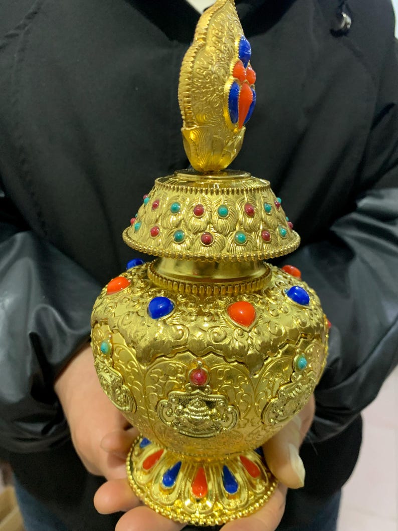 May include: Ornate gold-colored decorative vessel with a bulbous body and a tiered lid. The vessel is adorned with colorful gemstones in red, blue, and turquoise. The design features intricate patterns and a detailed finial.