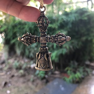 May include: An antique brass-coloured pendant in the shape of a cross. The pendant has ornate details, including floral and geometric patterns. It is suspended from a gold-coloured chain, and is held up by a finger.