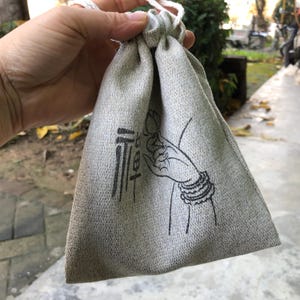 May include: A light gray drawstring bag with a black illustration of a hand holding a flower and text. The bag is made of a textured fabric and has a white drawstring closure. The illustration features intricate details.