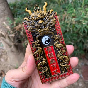 Könnte beinhalten: Ein rechteckiger, verzierter Anhänger mit einem bronzenen Drachenkopf oben. Der Anhänger hat einen roten Rand mit goldenen Drachen und chinesischen Schriftzeichen. Ein Yin-Yang-Symbol ist unter dem Drachenkopf zentriert.
