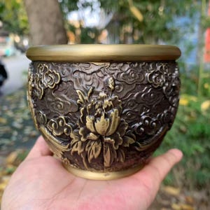 May include: A bronze-colored decorative bowl with a floral and cloud design. The bowl has a gold-colored rim and a detailed relief pattern of flowers and clouds. The bowl is held in a hand.