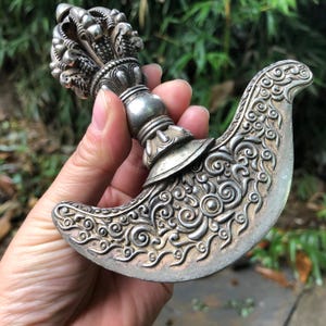 May include: A silver ritual knife with detailed floral and geometric patterns. The handle has an ornate design, and the curved blade is embellished with swirling motifs. The knife is held against a blurred green background.