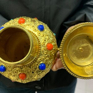 May include: Ornate gold-colored container with a lid, adorned with red and blue gemstones. The container has a detailed, embossed design and is open, revealing its interior. The lid is also gold and open, showing the inside of the lid.