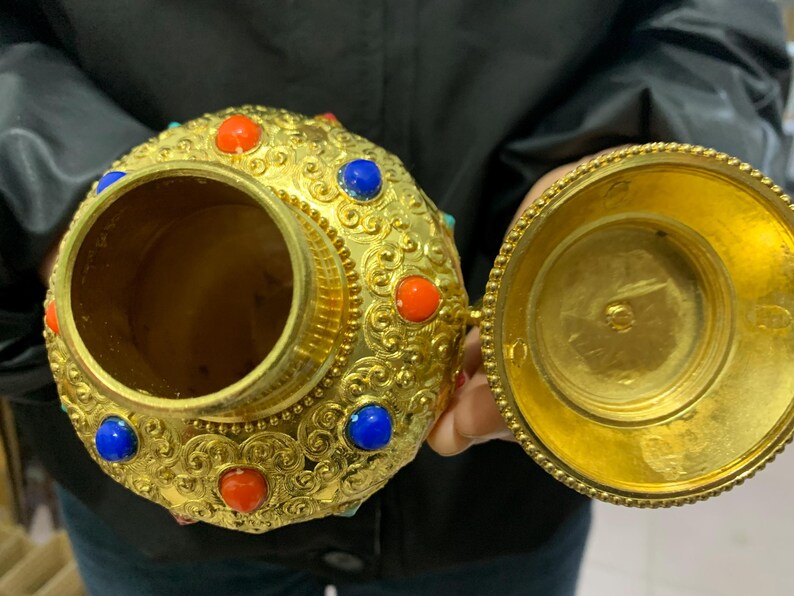 May include: Ornate gold-colored container with a lid, adorned with red and blue gemstones. The container has a detailed, embossed design and is open, revealing its interior. The lid is also gold and open, showing the inside of the lid.