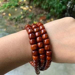 Op de afbeelding: Drie houten kralenarmbanden in een warme roodbruine tint. De kralen zijn glad en gepolijst. De armbanden zijn op elkaar gestapeld, waarbij de natuurlijke houtnerf zichtbaar is. Elke armband heeft een omtrek van ongeveer 20 cm.