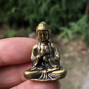 Op de afbeelding: Een klein, antiek ogend messing Boeddha-beeld in een zittende meditatiehouding. De figuur heeft een serene uitdrukking en is gedetailleerd met traditionele gewaden en een patroonhoofddeksel. Het beeld wordt in een hand gehouden tegen een vage groene achtergrond.