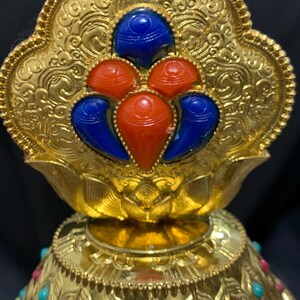 May include: Ornate gold-colored decorative object with intricate patterns and embedded gemstones. The top features a floral design with blue and red stones. The base is adorned with turquoise and red stones, creating a striking contrast against the gold.