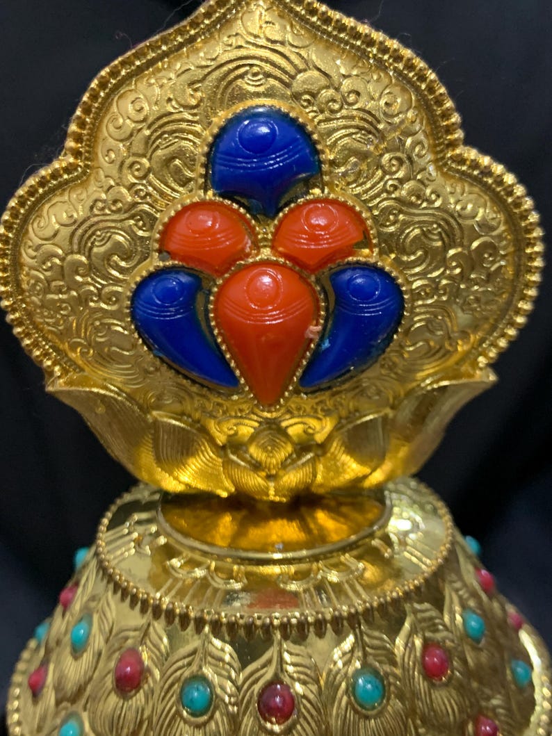 May include: Ornate gold-colored decorative object with intricate patterns and embedded gemstones. The top features a floral design with blue and red stones. The base is adorned with turquoise and red stones, creating a striking contrast against the gold.