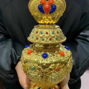 May include: Ornate gold-colored decorative vessel with intricate embossed patterns and colorful gemstone accents. The vessel features red, blue, and turquoise stones, and a lotus-shaped top. The design is highly detailed and symmetrical.