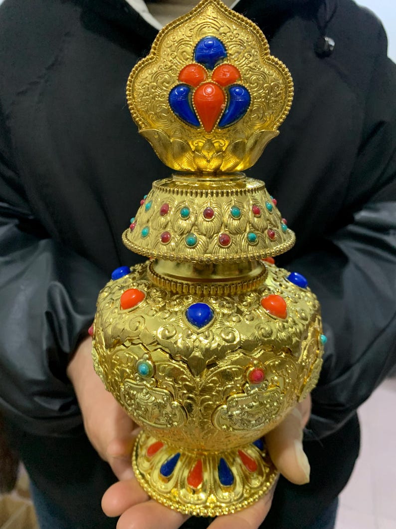 May include: Ornate gold-colored decorative vessel with intricate embossed patterns and colorful gemstone accents. The vessel features red, blue, and turquoise stones, and a lotus-shaped top. The design is highly detailed and symmetrical.