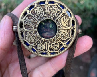 May include: A circular pendant in an antique style, with a gold-coloured metal frame and detailed embossed patterns. The pendant has a central image and blue accents. A brown leather cord is attached for wearing.
