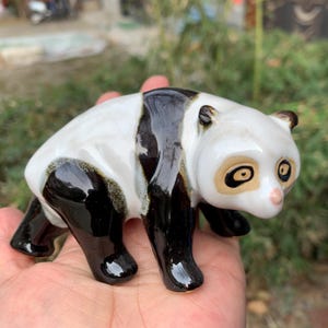 May include: A ceramic panda figurine with a glossy finish. The panda has a white body with black markings on its legs, back, and ears. The panda's face has tan accents around the eyes and a pink nose. The figurine is in a crouching position.