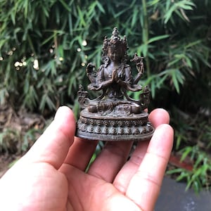 Puede incluir: Pequeña estatua de color bronce de una deidad con múltiples brazos y una corona. La figura está sentada con las manos juntas en oración. La estatua está sobre una base decorativa, sostenida en la mano de una persona contra un fondo verde borroso.