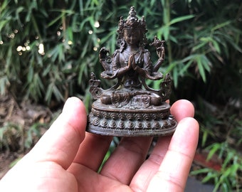 May include: A small, bronze-coloured statue of a deity with multiple arms and a crown. The figure is seated with hands clasped in prayer. The statue is on a decorative base, held in a person's hand against a blurred green background.