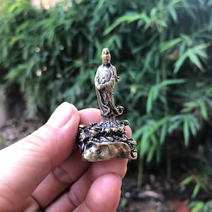 Può includere: Una piccola statua color bronzo di una figura in piedi con abiti fluenti. La figura ha un'espressione serena ed è su una base dettagliata. La statua è tenuta in mano su uno sfondo di fogliame verde.