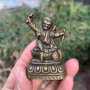 May include: A bronze-colored figurine of a seated deity, holding a snake and a small weapon. The figure is atop a detailed base with a floral design and inscribed text. The statue is held in a hand against a blurred green background.
