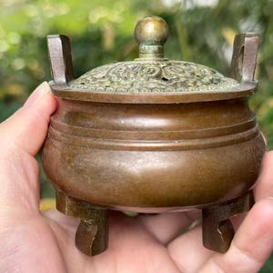 May include: A small, antique bronze incense burner with a decorative lid. The burner has a round body with a detailed, pierced lid and three small feet. The lid is topped with a small, round knob. The overall color is a warm, aged bronze.