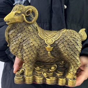 May include: A detailed brass sculpture of a ram, featuring intricate coin-like scales and curled horns. The ram has a saddle-like design on its back and is standing on a base with several small bowls. The sculpture is a golden color.